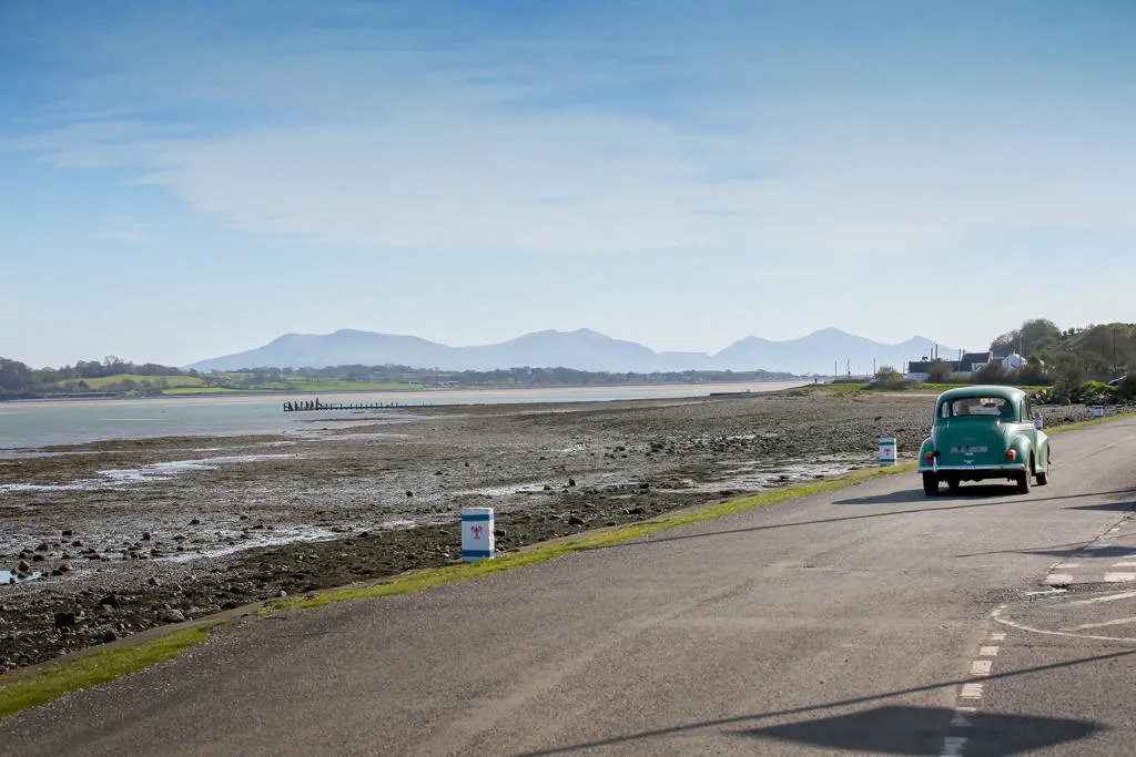Old Morris Minor car on road by entry to the Sea Zoo and Halen Môn, with the remains of the Foel Ferry Pier to the left