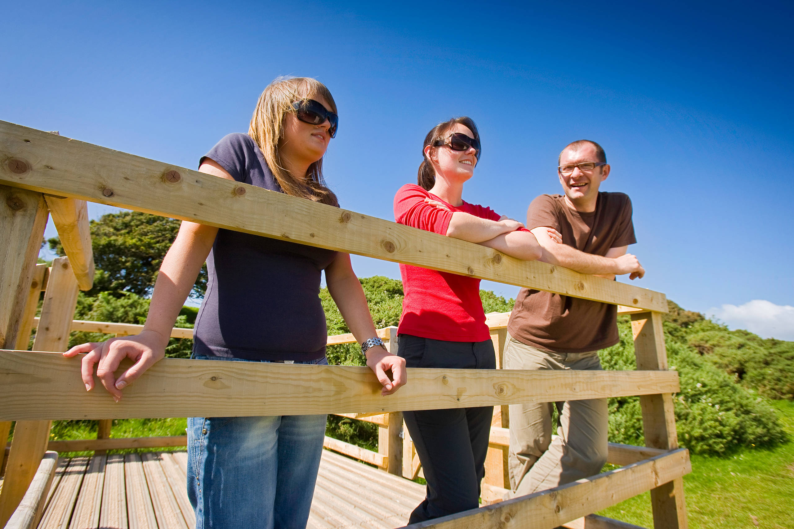 Three people on the board walk looking over Llyn Maelog on a Sunny day