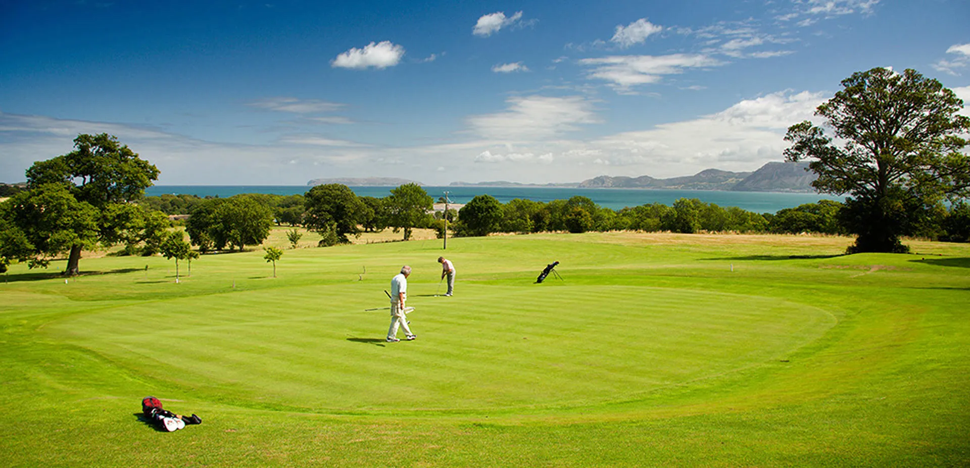 Two people playing golf on the final green trying to putt with golf bag lying on floor