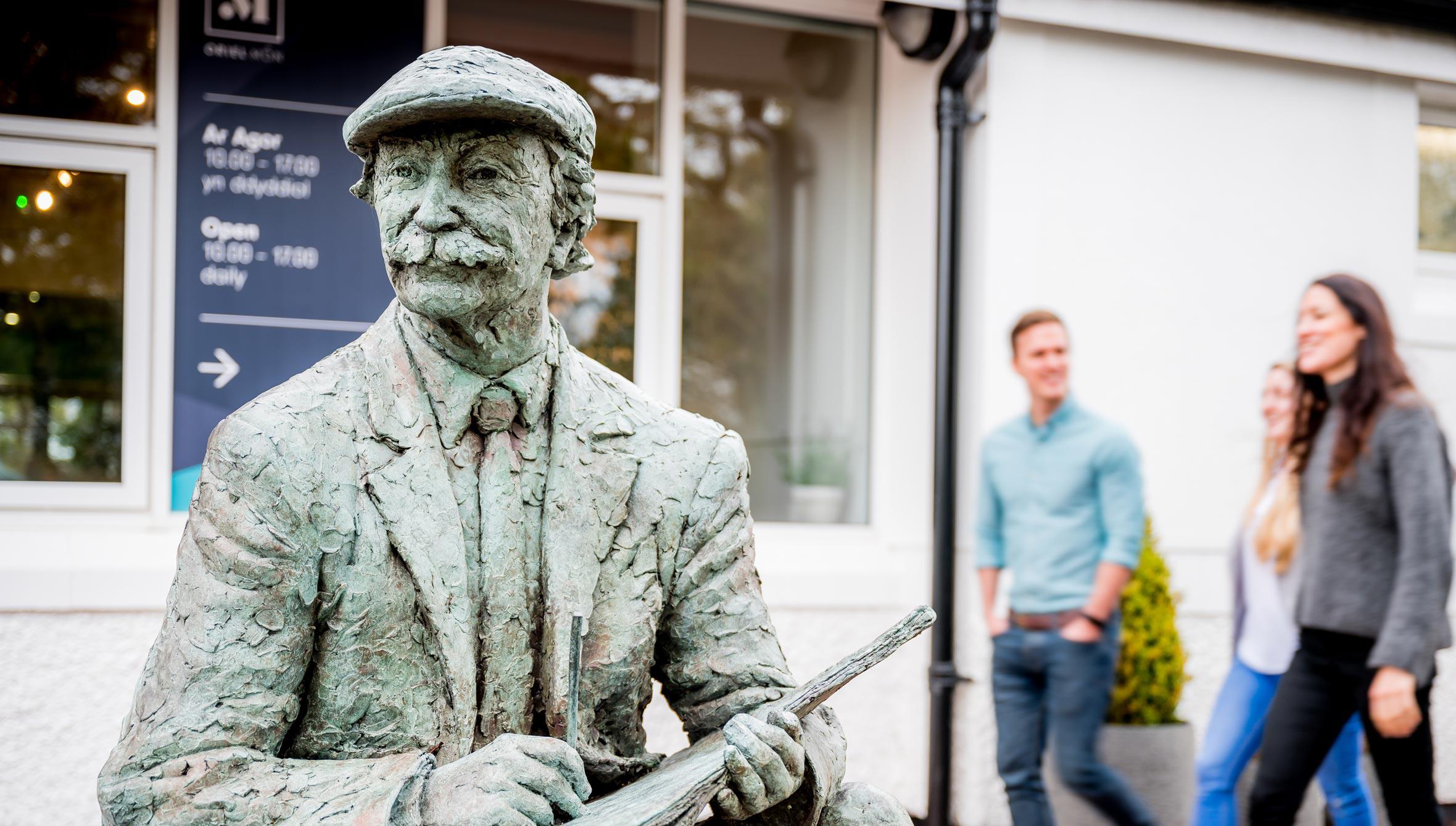 Statue of the artist Kyffin Williams outside Oriel Môn with people walking in the background